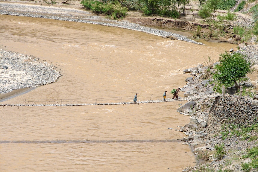 A man and a boy carry a load on their backs across a rickity bridge.