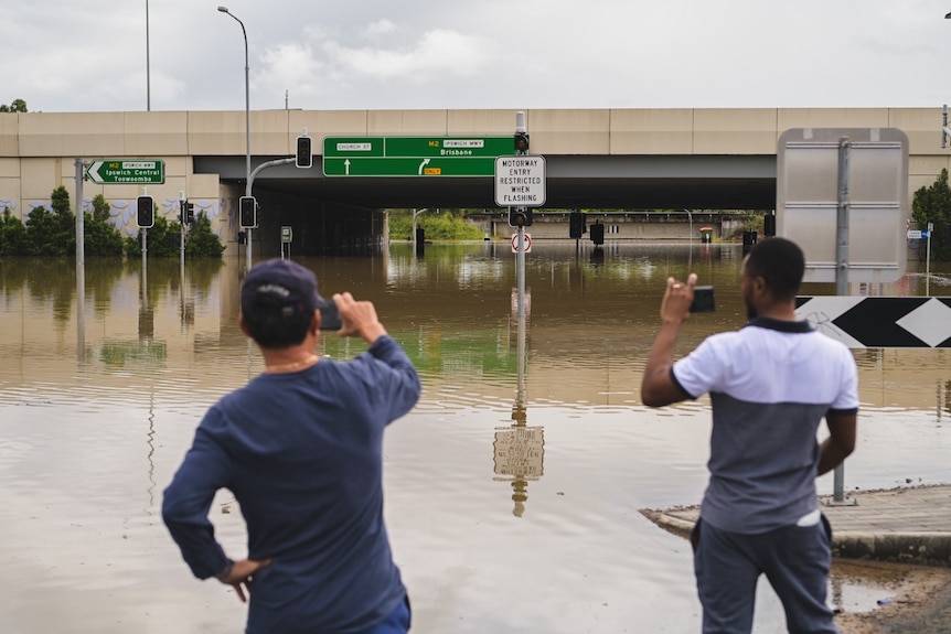 Two men stand with their backs to the camera, taking photos of brown floodwaters that have covered a busy intersection.