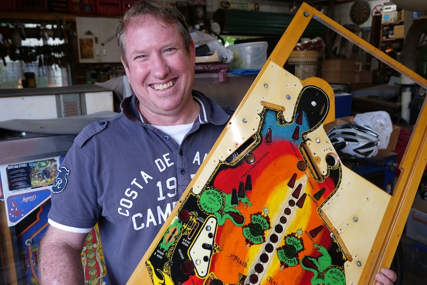 Lloyd smiles proudly holding the base for the Black Knight pinball machine, with bright artwork.