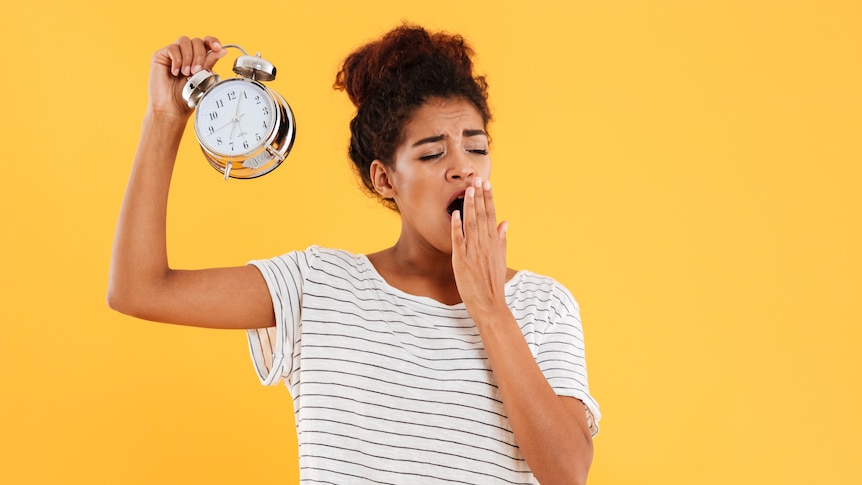 A tired young African woman yawns holding alarm clock