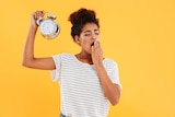 A tired young African woman yawns holding alarm clock