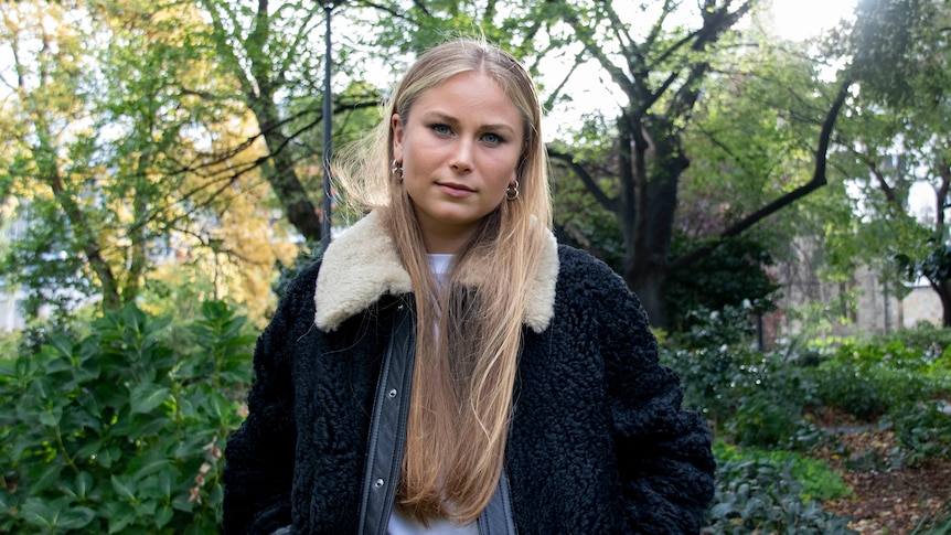 A young woman with long blonde hair standing in a park looking at the camera.