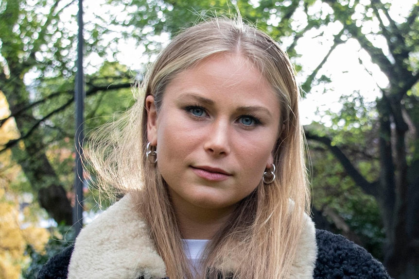 A young woman with long blonde hair standing in a park looking at the camera.