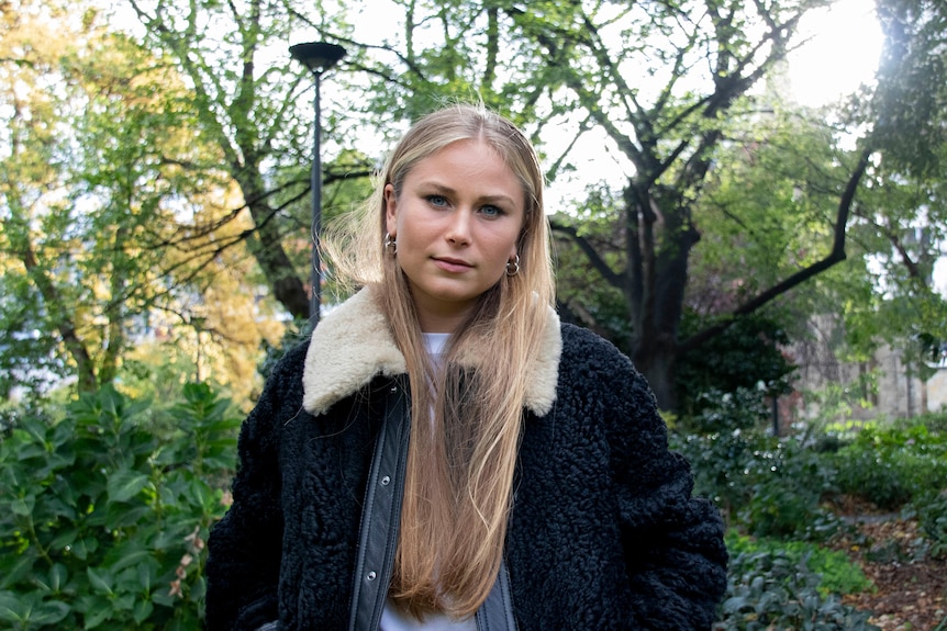 A young woman with long blonde hair standing in a park looking at the camera.