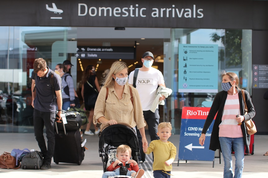 Passengers with bags and face masks, including a woman with two small children, walk out of the exit at Perth Airport.