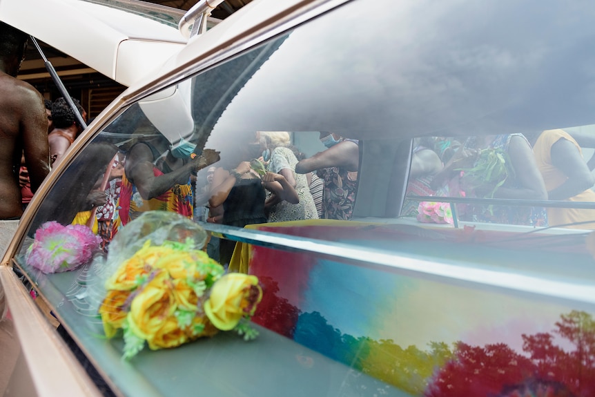 A coffin in the back of a hearse is surrounded by people.