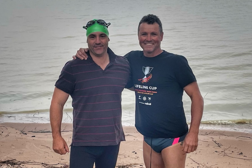 Two men in swimming attire standing on a lake shore in front of a large body of water