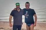 Two men in swimming attire standing on a lake shore in front of a large body of water
