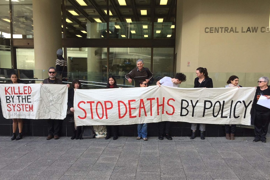 Protestors hold up signs saying "Stop deaths by custody" and "killed by the system" with a crane on it.