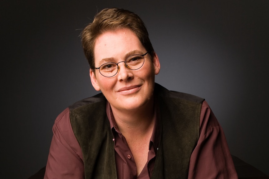 A woman with short hair and glasses sits in front of a black background.