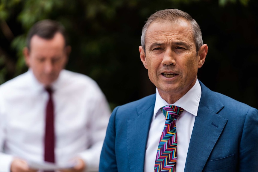 Man in blue suit and tie, head and shoulders shot with another man blurred in the background