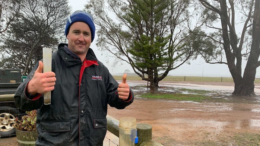 A farmer wearing a beanie and a jacket stands outside holding a rain gauge.