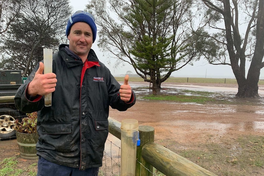 A farmer wearing a beanie and a jacket stands outside holding a rain gauge.