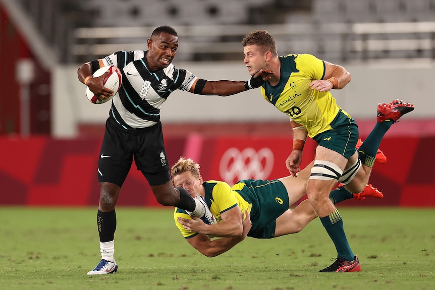 A fiji player in white and black is tackled by two australian players in yellow