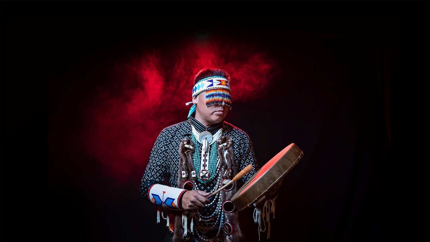 In front of red smoke, a man beats drum with beaded head piece covering eyes and wears intricate native American regalia.