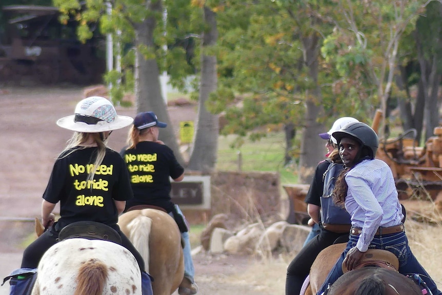Horses and riders walking away from camera with t-shirts saying "no need to ride alone"