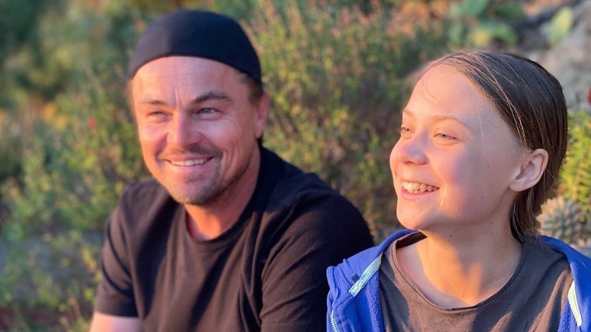 Leonardo DiCaprio and Greta Thunberg smile together while sitting outside among greenery.