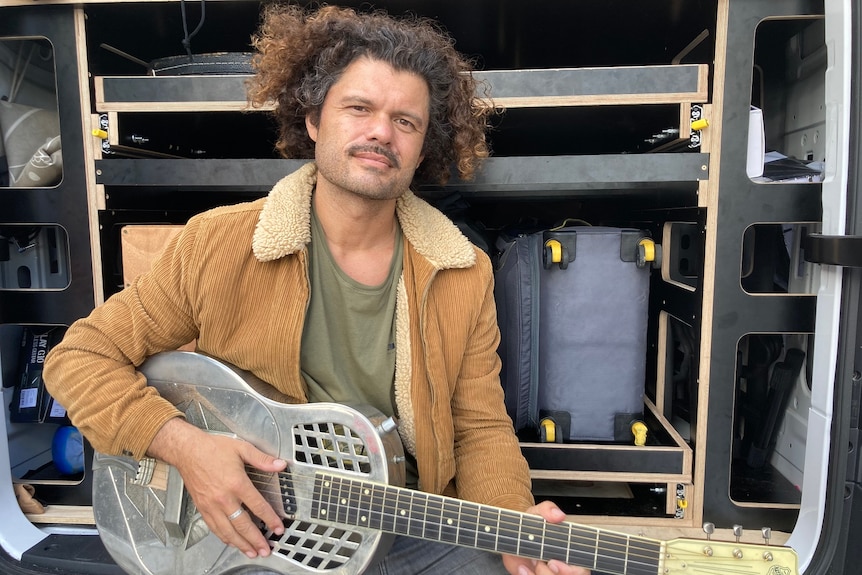 A young man with curly hair poses with his guitar at the back of his van.