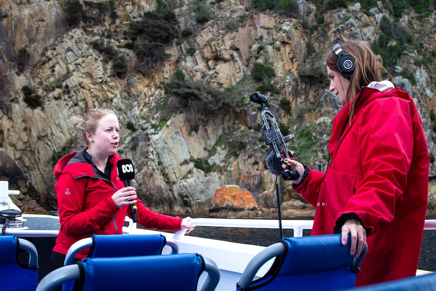 Two women stand on a boat in the cold, while one films the other holding a microphone.