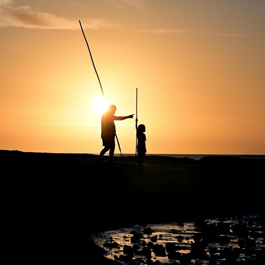 The silhouette of a father and son going to hunt seafood using traditional spears, set against a golden sunset.