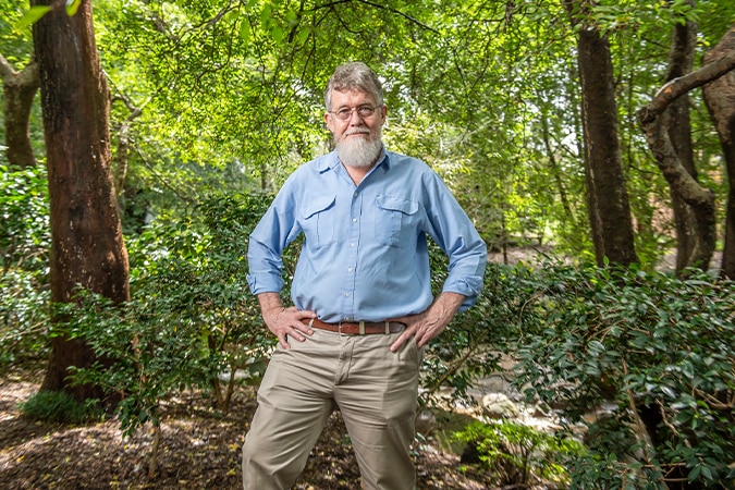 A man wearing chinos and a button-up shirt stands in a forest clearing with his hands on his hips.