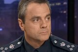 Stuart Bateson, wearing a police uniform, looks to his left while sitting on the set of Q&A