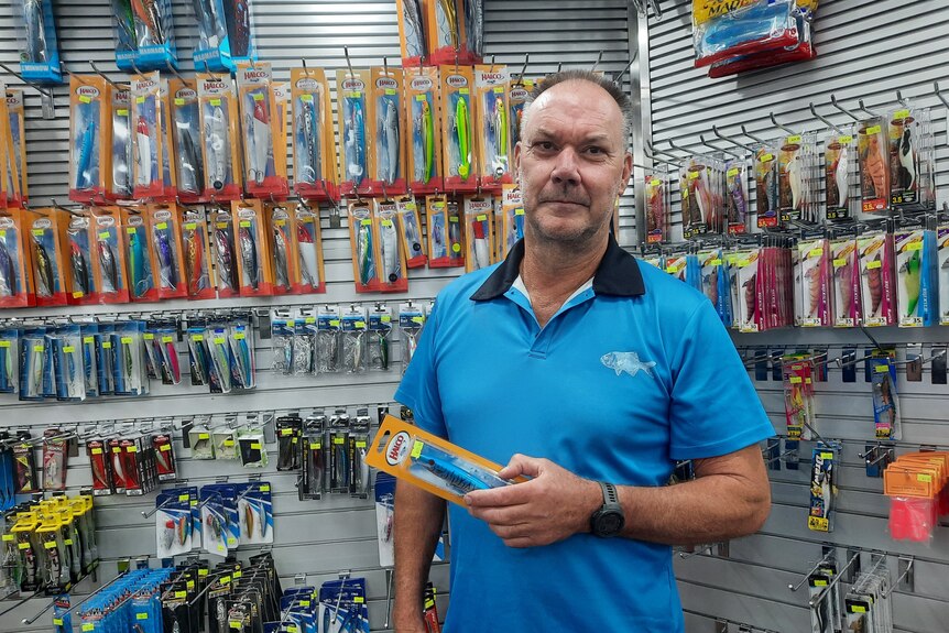 Man in blue shirt stands in tackle shop in front of fishing gear on a wall.