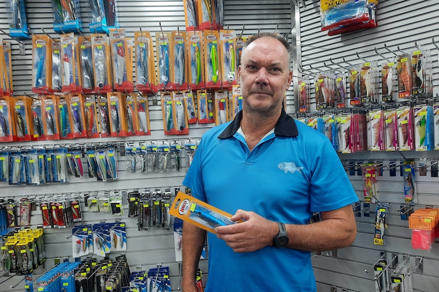 Man in blue shirt stands in tackle shop in front of fishing gear on a wall.