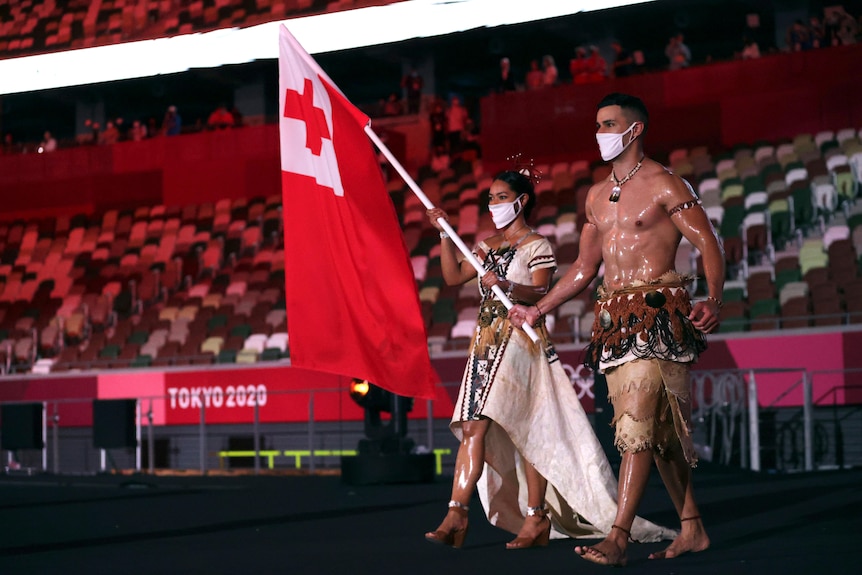 A male and female flagbearer from tonga walk into olympic stadium in japan. the male flagbearer is not wearing a shirt