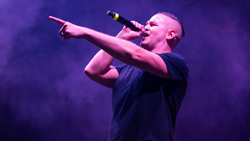 Suffa from Hilltop Hoods stands on stage, pointing into the distance whilst holding a microphone to his mouth