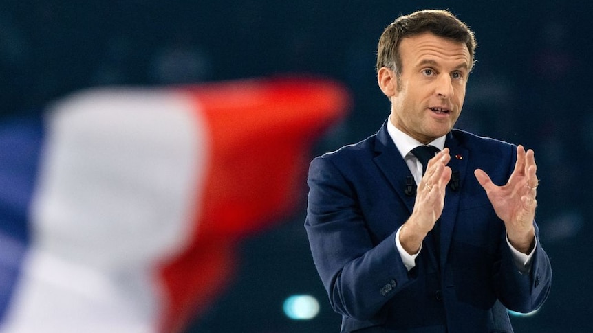 French President Macron with a blurred french Flag behind him and hands pointing forward
