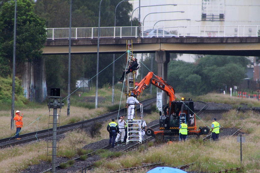 Emergency services stand around a man on attached to a pole over a railway track