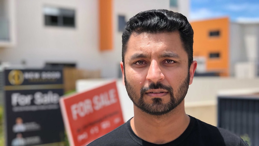Najam looks into the camera, standing in front of some 'for sale' signs outside his building.