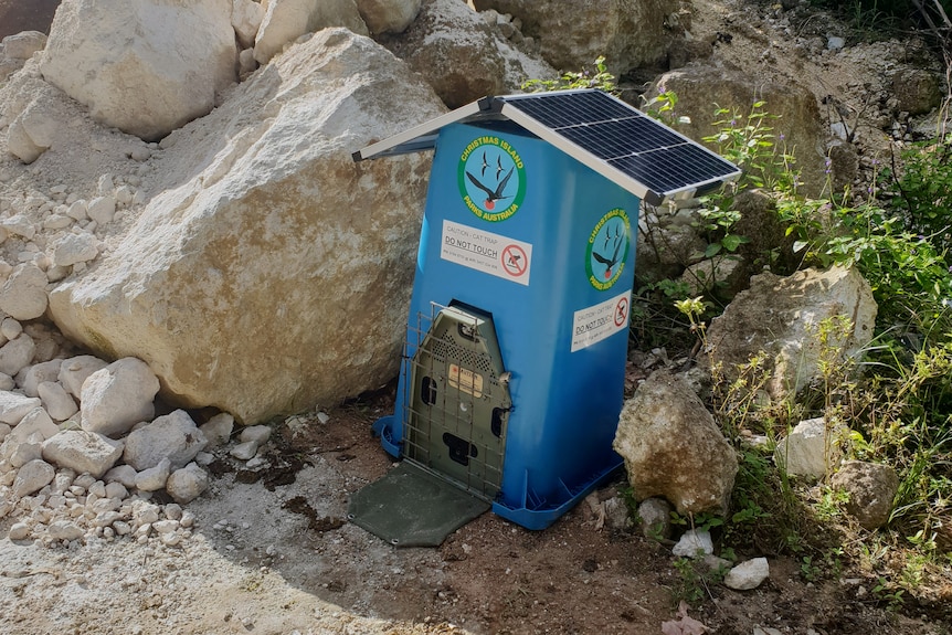 A silver device with a cage around it with an upsidedown blue wheelie bin on top with solar panel slats in a rocky outcrop.