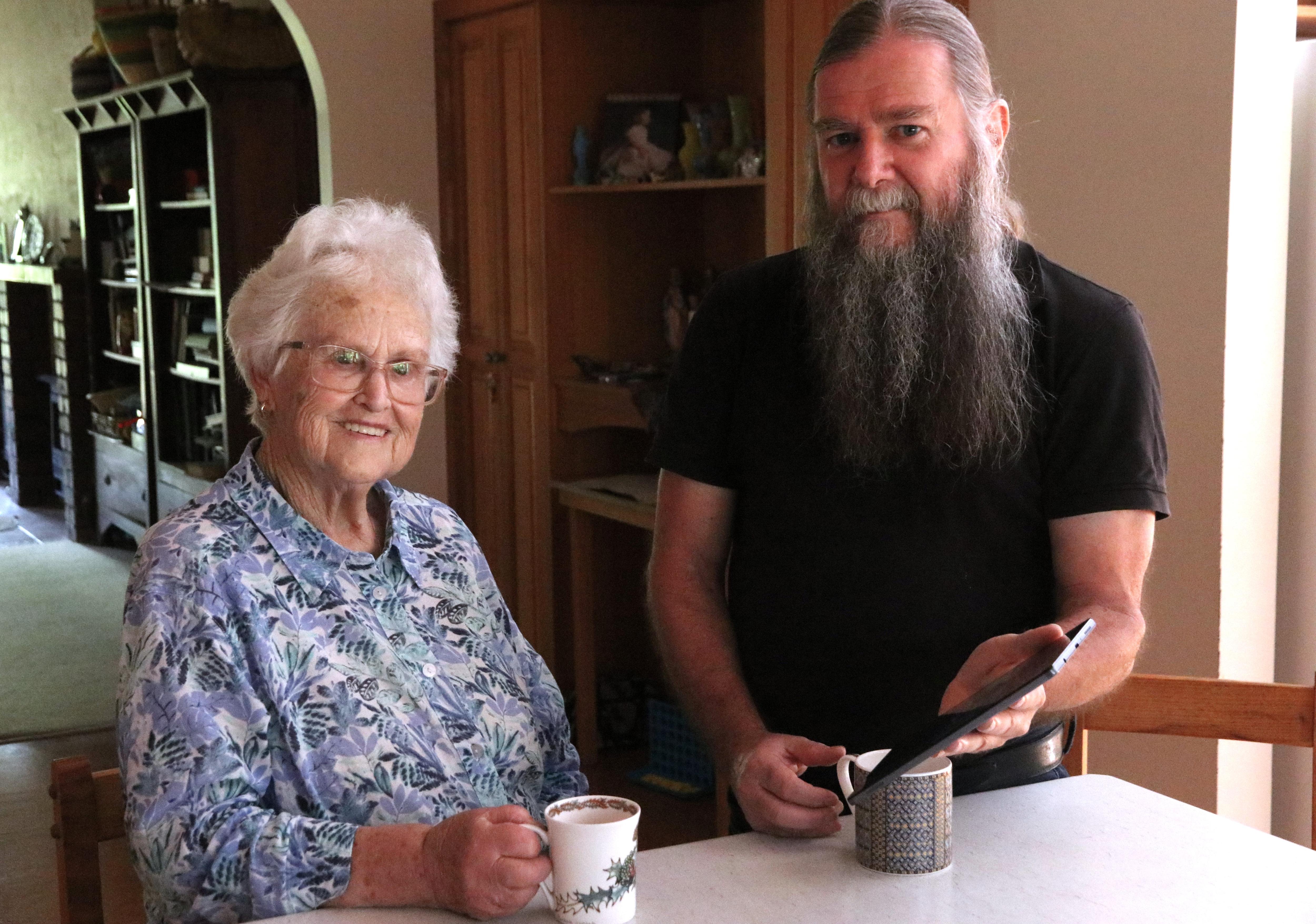 Elderly lady and man with a beard, sitting at bench with a cup of tea and ipad, looking at camera 