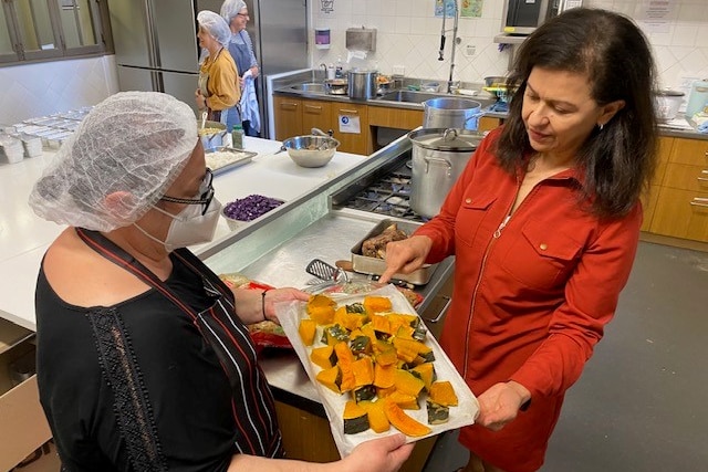 A woman in a red dress looks at a platter of roasted kent pumpkin held by a volunteer working in a commercial kitchen.