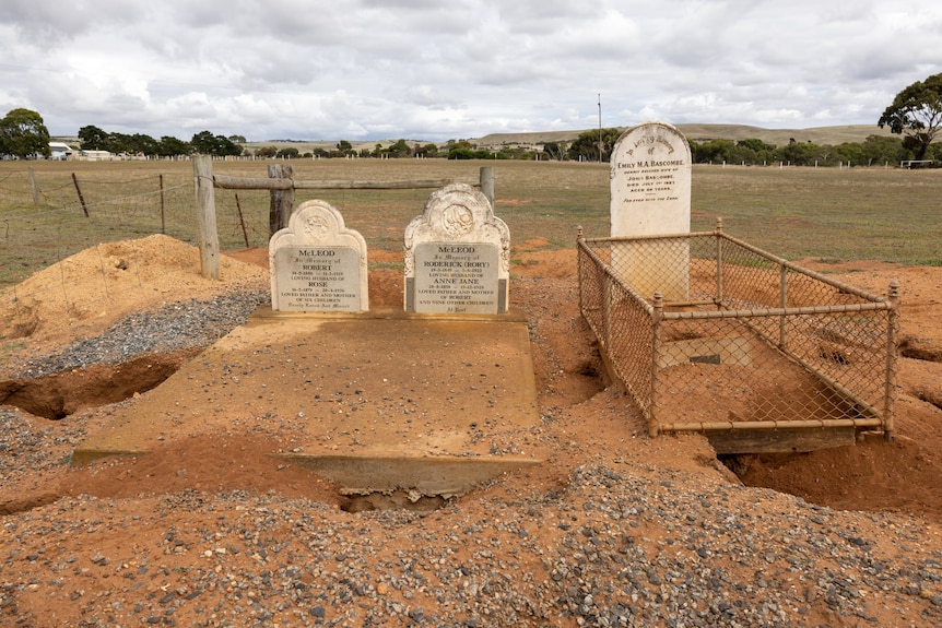 A photo of three headstones with the ground in front of them eroded, uneven and full of holes and dips.