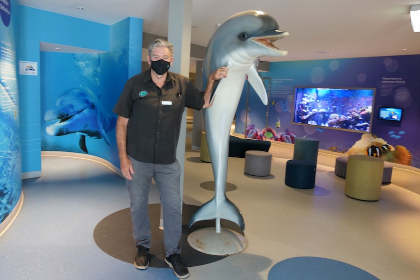 Man wearing COVID mask stands next to dolphin statue in aquarium.