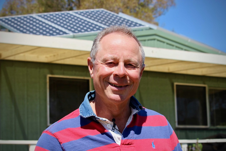 A man with short grey hair stands in front of a house with solar panels on the roof.