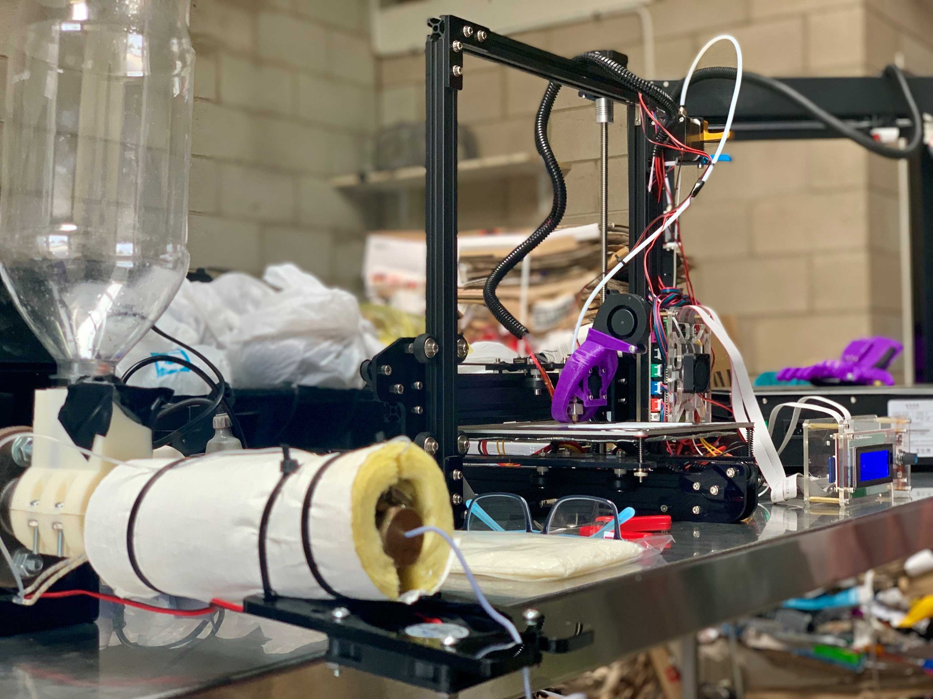 Una pequeña extrusora de plástico se sienta en un banco junto a una impresora 3D