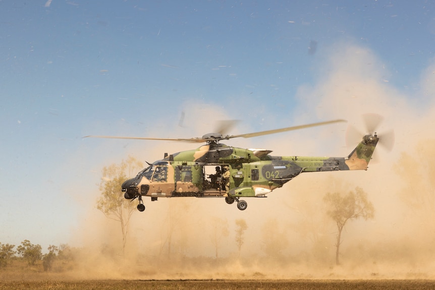 A military helicopter lands in the bush, kicking up clouds of dust.