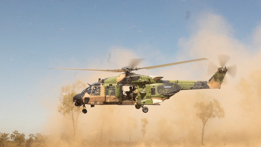 A military helicopter lands in bushland, kicking up clouds of dust.