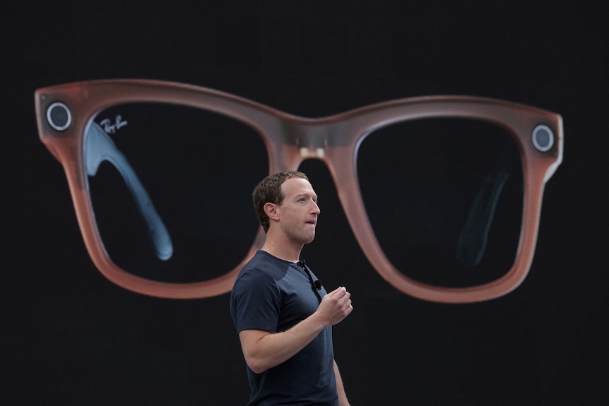 A white man stands and delivers a speech in front of a pair of smart sunglasses being displayed on a large screen behind him.