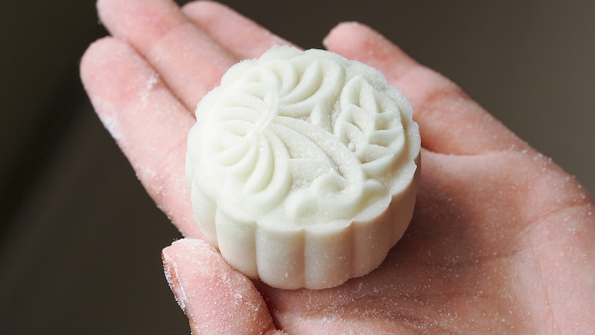 A person's palm, holding a white round mooncake with a floral decoration embossed into it.