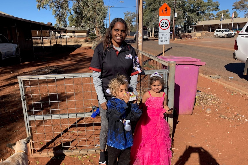 A smiling staff member stands behind two children on a red sandy road with a pink bin