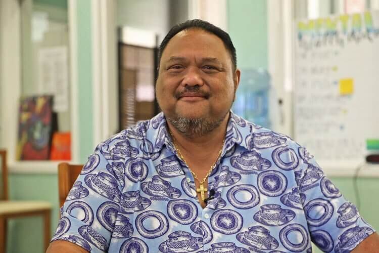 Mark Kawika Patterson, wearing brightly coloured print shirt and a gold crucifix necklace, sits smiling.