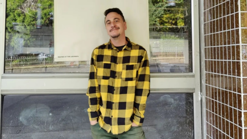 A young man in a bright, yellow check shirt stands in front of a shop.