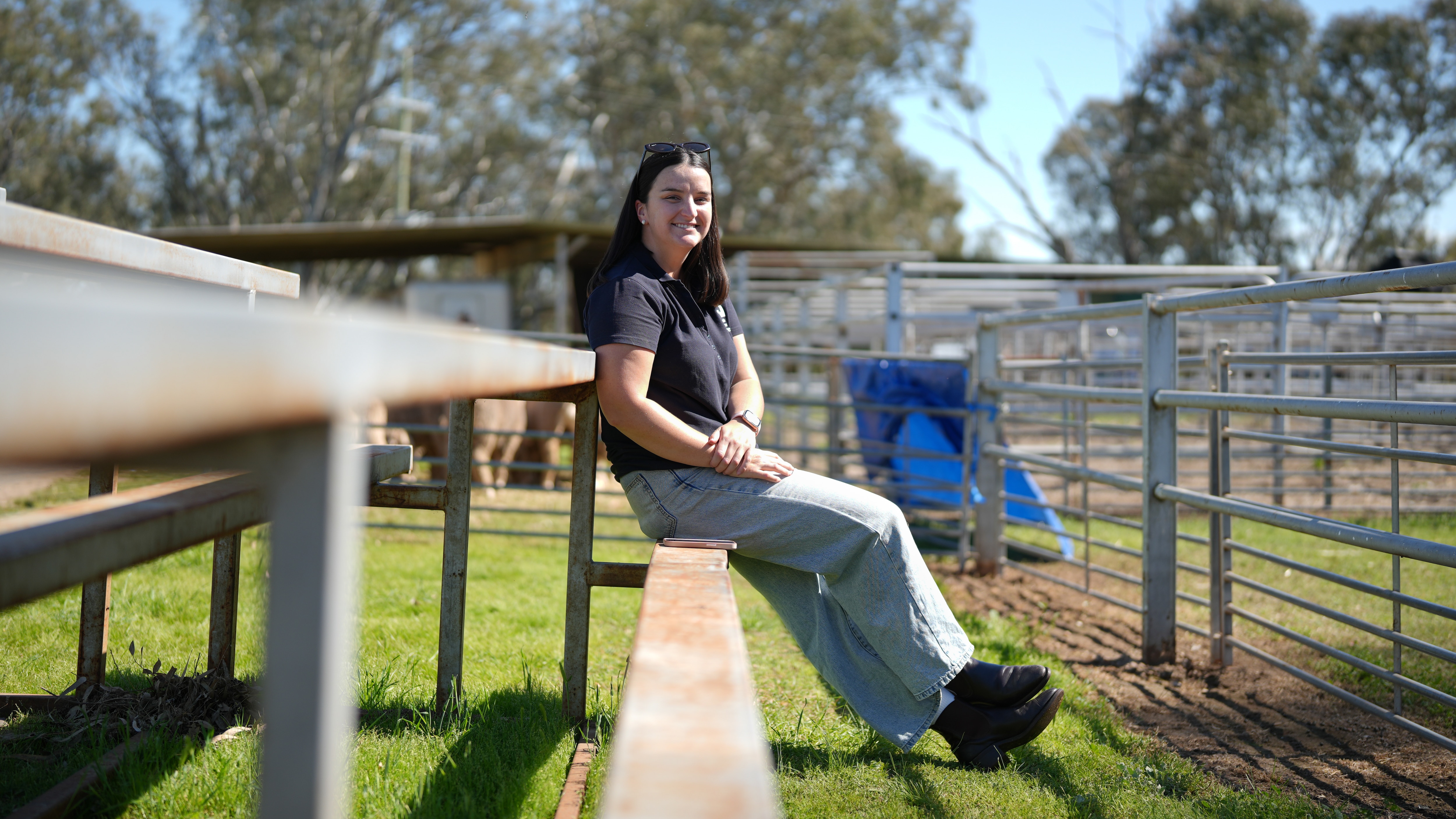Woman smiling sitting on a metal bench in a paddock with a sheep pen.