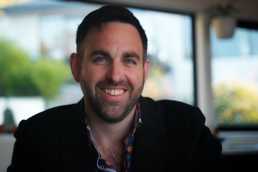 A man with dark hair and blue eyes in a black jacket smiles at the camera.