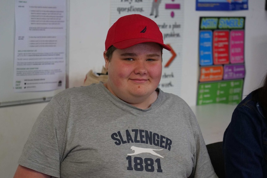A teenage boy in a classroom wearing a t-shirt and a red cap.